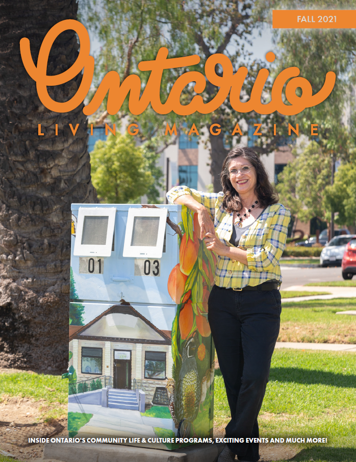 Female artist in yellow plaid shirt, stands smiling proudly, arm on top of painted utility box. 