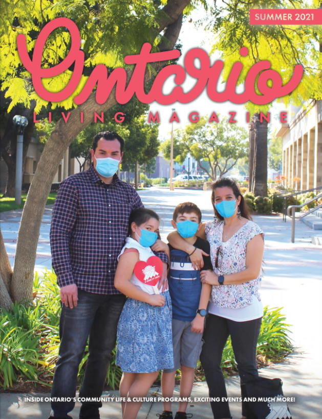 Family standing close together in front of bright, sun-lit trees, looking straight forward at the camera while wearing face masks.