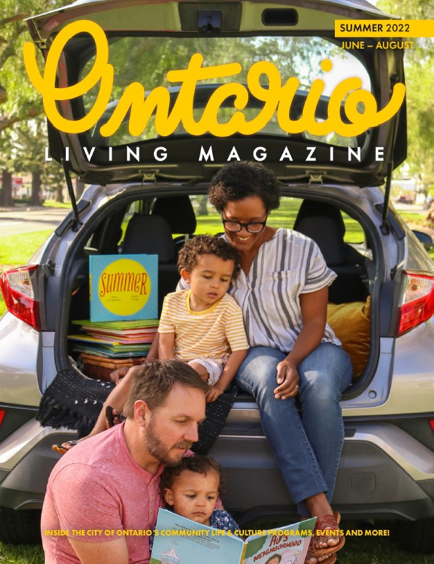 Local author sits in the open trunk of a hatchback with their family, smiling, reading a book. 